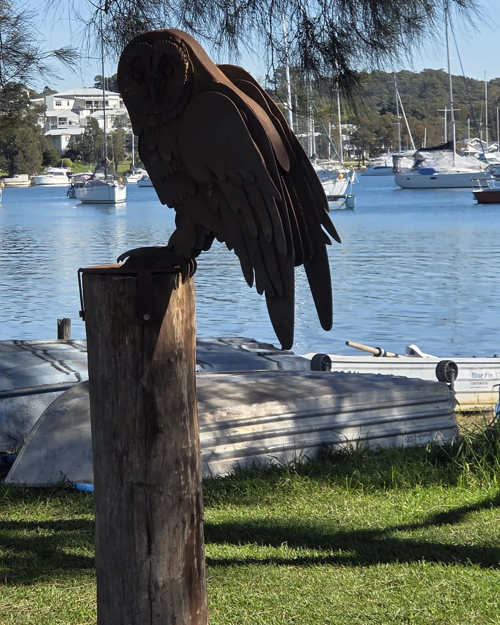 Rusted steel owl sculpture installed on a timber post beside the water, boats moored behind